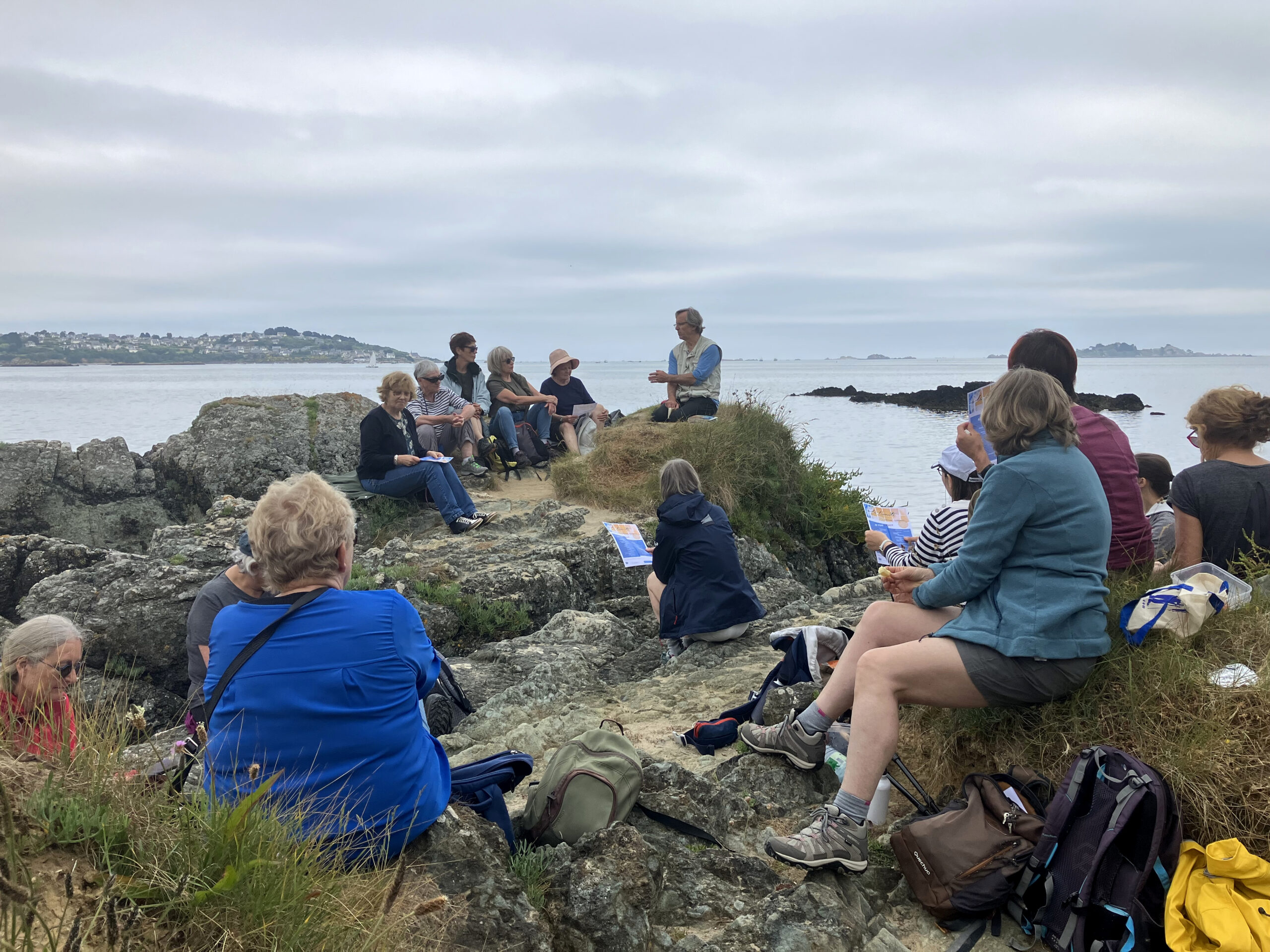 Un groupe assis sur des rochers devant la mer écoute Marc Roger lire des textes.